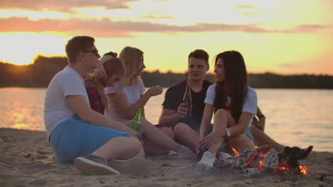 The-group-of-students-is-sitting-in-around-bonfire-on-the-lake-coast.-They-are-spending-time-cheerfully-and-they-are-talking-to-each-other-and-drinking-beer-at-sunset.
